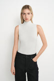 A blonde model wearing a sleeveless, high-neck white top and dark wash jeans, standing against a plain white studio background.