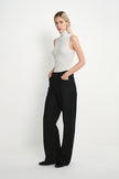 A blonde model wearing a white sleeveless mock-neck top and black wide-leg trousers, standing against a plain white studio background.