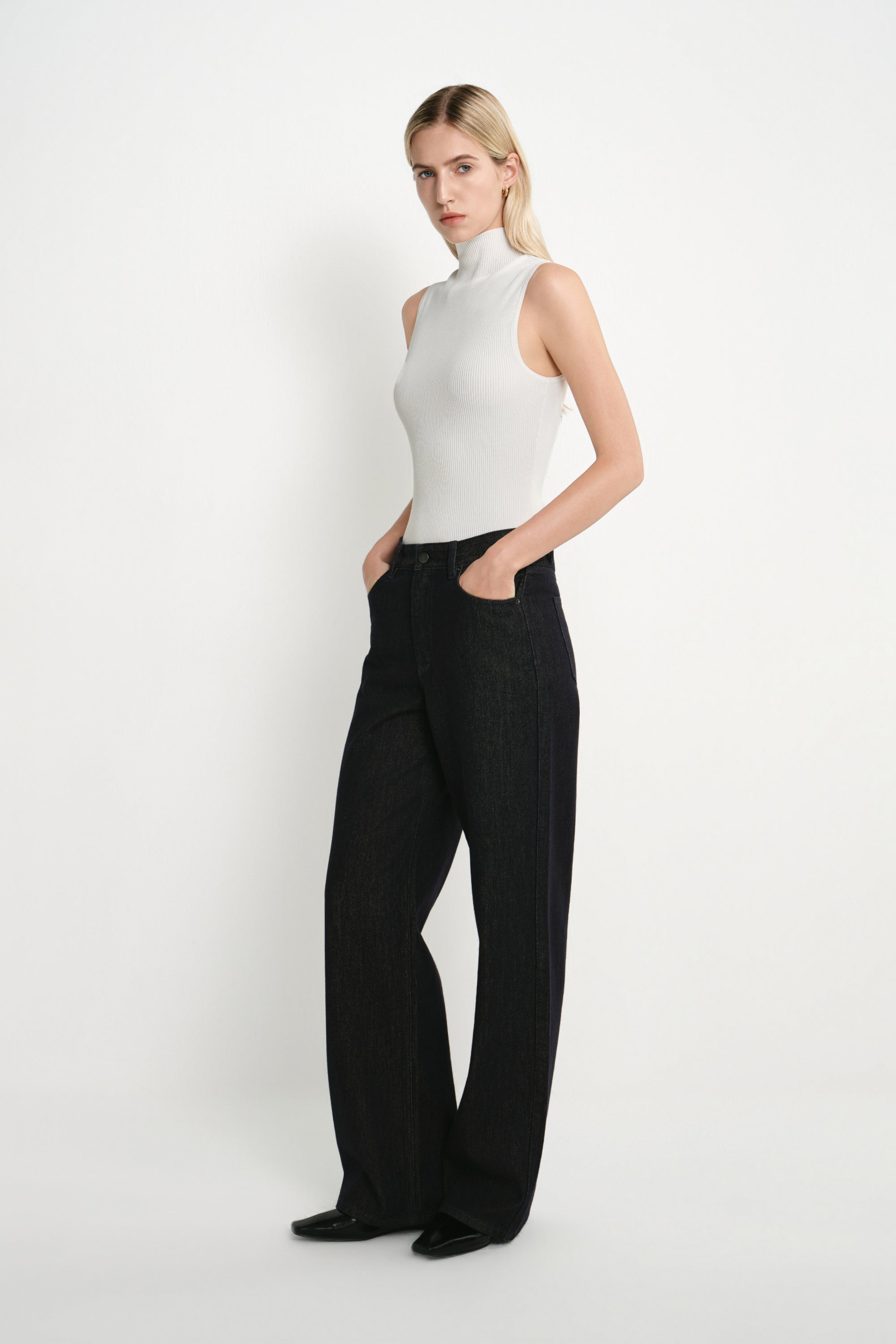 A blonde model wearing a white sleeveless mock-neck top and black wide-leg trousers, standing against a plain white studio background.