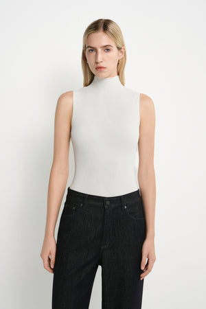 A blonde model wearing a fitted, sleeveless white mock-neck top and dark wash jeans, standing against a plain white studio background.