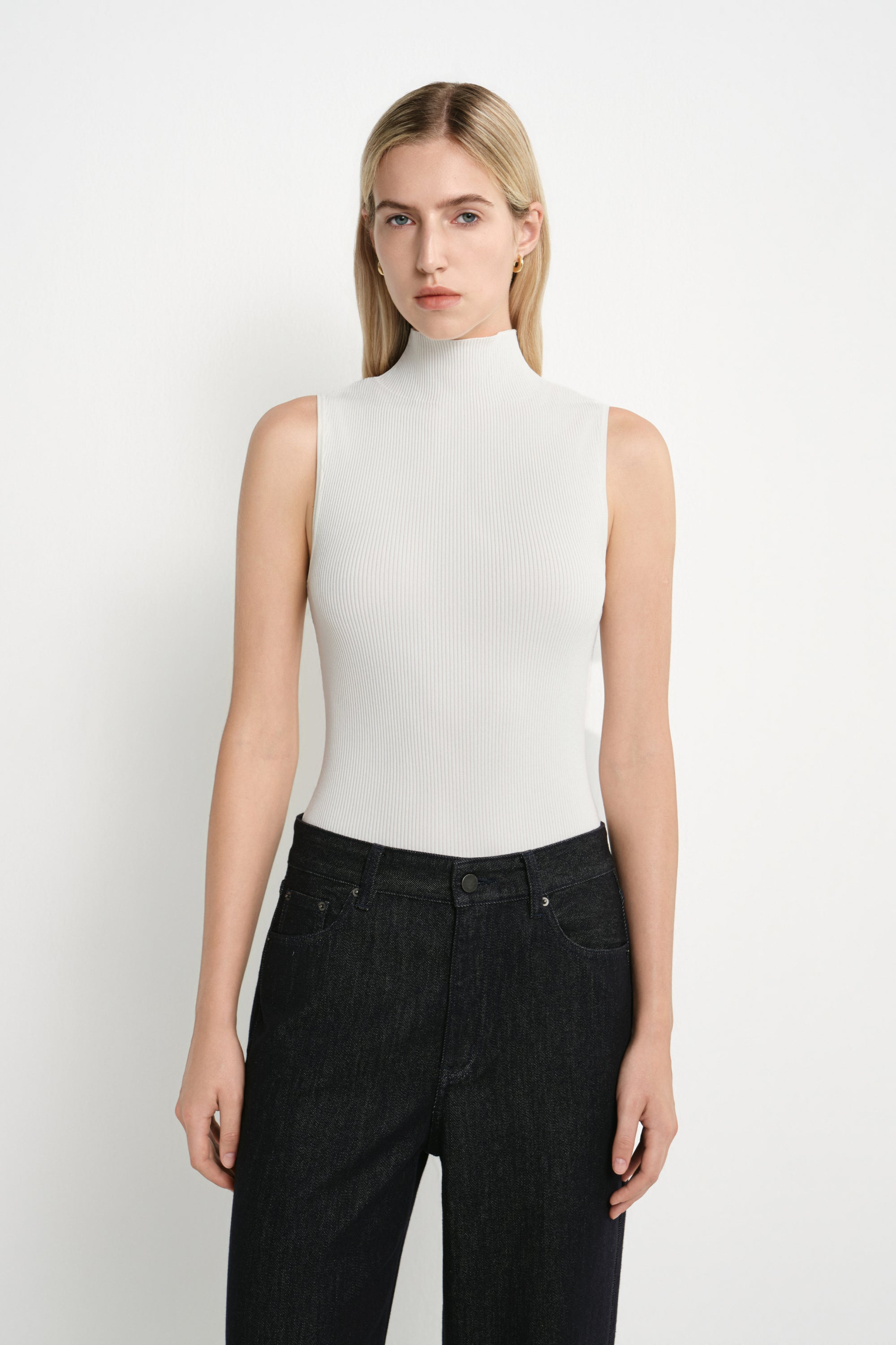 A blonde model wearing a fitted, sleeveless white mock-neck top and dark wash jeans, standing against a plain white studio background.