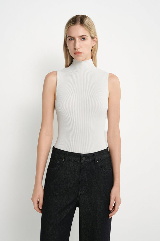 A blonde model wearing a fitted, sleeveless white mock-neck top and dark wash jeans, standing against a plain white studio background.