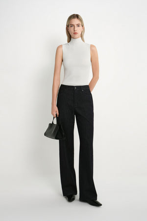 A model stands against a plain white studio background wearing a sleeveless white mock-neck top, black wide-leg jeans, black pointed shoes, and holding a small black handbag.