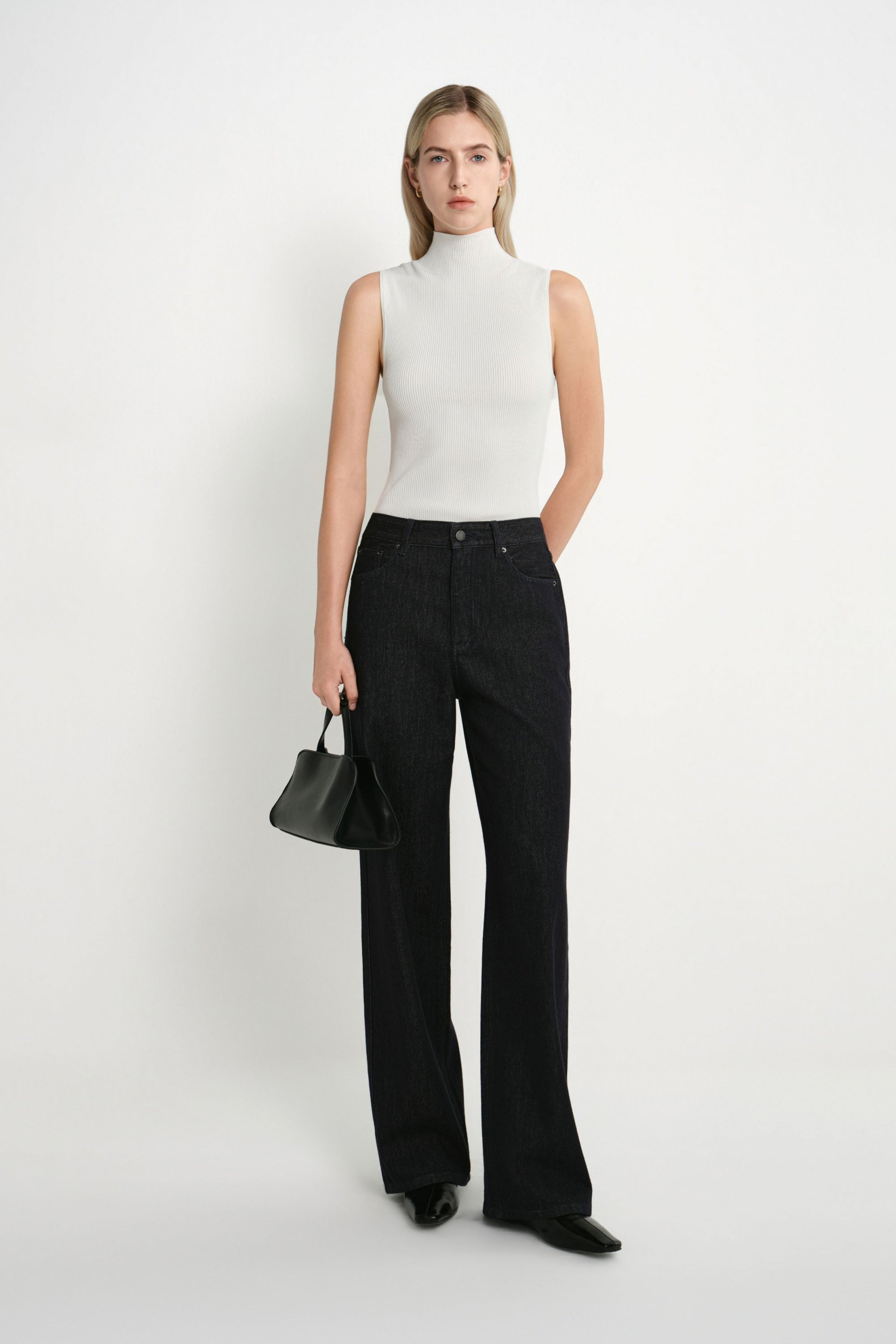 A model stands against a plain white studio background wearing a sleeveless white mock-neck top, black wide-leg jeans, black pointed shoes, and holding a small black handbag.