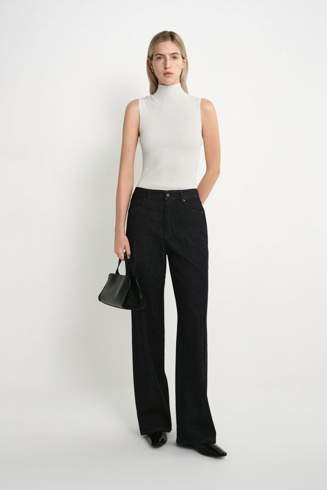 A model stands against a plain white studio background wearing a sleeveless white mock-neck top, black wide-leg jeans, black pointed shoes, and holding a small black handbag.