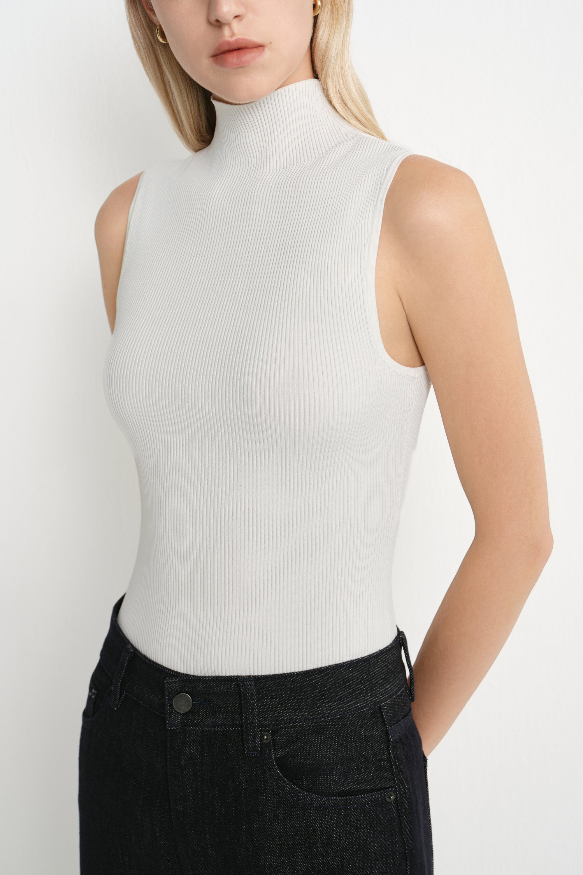 A woman wearing a ribbed sleeveless mock neck top in off-white, paired with dark denim jeans, shown from the chest up against a plain light background.