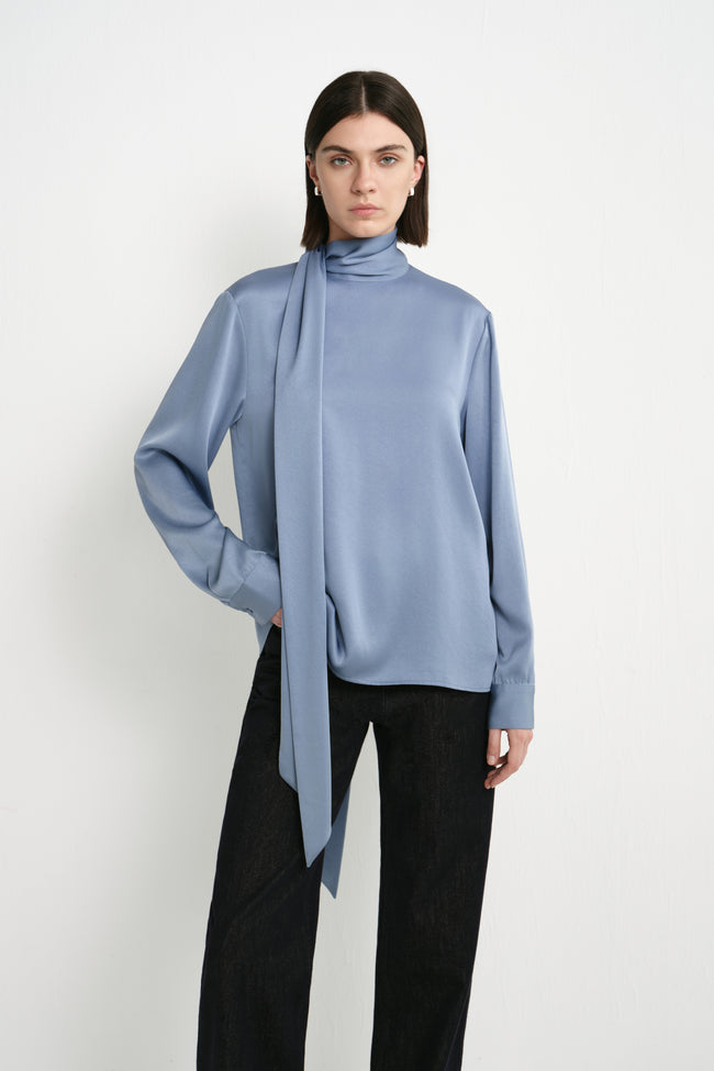 A model wearing the Rue Sophie Samara Tie Neck Top in Harbour with a high scarf neckline, elongated tie detail, buttoned cuffs, and a softly draped silhouette, front view.
