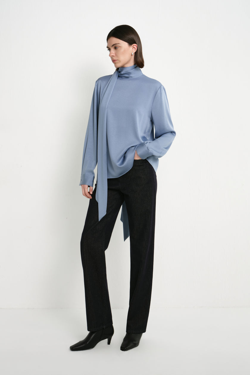 A model wearing the Rue Sophie Samara Tie Neck Top in Harbour with a high draped neckline, long scarf tie, and relaxed flowing silhouette, three-quarter front view.
