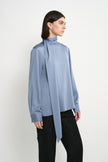 A model wearing the Rue Sophie Samara Tie Neck Top in Harbour, highlighting the high scarf neckline, elongated tie detail, long sleeves, and smooth draped finish, three-quarter view.
