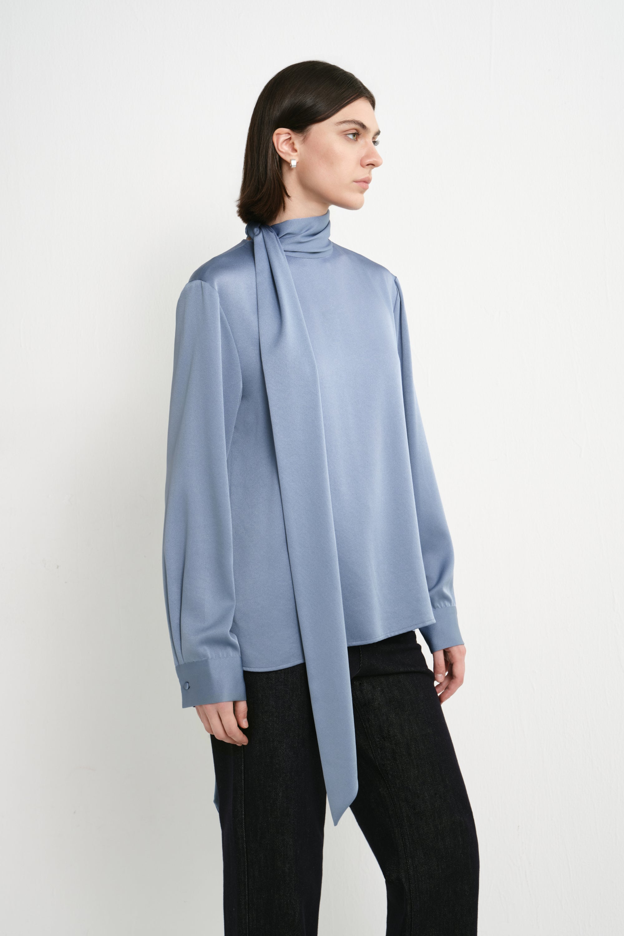 A model wearing the Rue Sophie Samara Tie Neck Top in Harbour, highlighting the high scarf neckline, elongated tie detail, long sleeves, and smooth draped finish, three-quarter view.