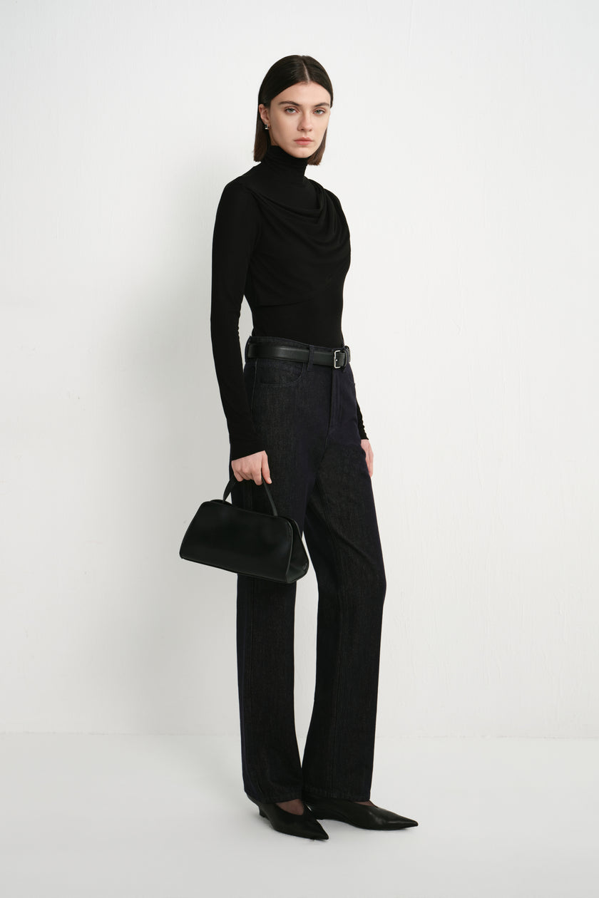 A model wearing Rue Sophie’s Lila Drape Scarf Jersey Top in Black with dark jeans and a black handbag, showing the fitted silhouette and asymmetric draped neckline.