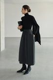 A model wearing Rue Sophie’s Lila Drape Scarf Jersey Top in Black with a grey drawstring midi skirt, showing the asymmetric shoulder drape in a styled editorial look.