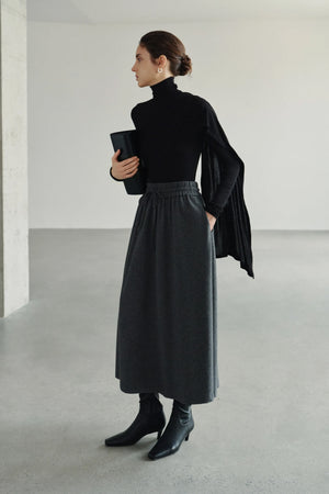 A model wearing Rue Sophie’s Lila Drape Scarf Jersey Top in Black with a grey drawstring midi skirt, showing the asymmetric shoulder drape in a styled editorial look.