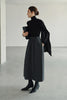 A model wearing Rue Sophie’s Lila Drape Scarf Jersey Top in Black with a grey drawstring midi skirt, showing the asymmetric shoulder drape in a styled editorial look.