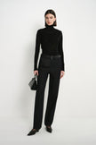 A model wearing Rue Sophie’s Lila Drape Scarf Jersey Top in Black with dark wide-leg jeans, highlighting its high neckline and asymmetric draped front.