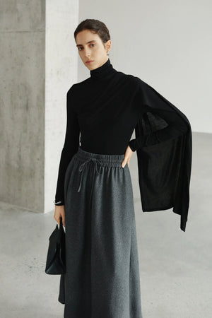 A model wearing Rue Sophie’s Lila Drape Scarf Jersey Top in Black with a grey drawstring skirt, highlighting the high neckline and long scarf-like drape extending from the shoulder.