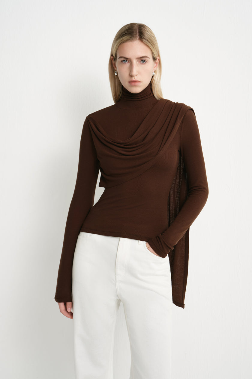 Front close-up of a dark brown fitted turtleneck top with soft layered draping across the chest and an asymmetric scarf-like panel over the shoulder.