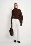 A model wearing a dark brown draped turtleneck top with a cape-like asymmetric overlay and long side panel, styled with white straight-leg jeans and a brown bag.