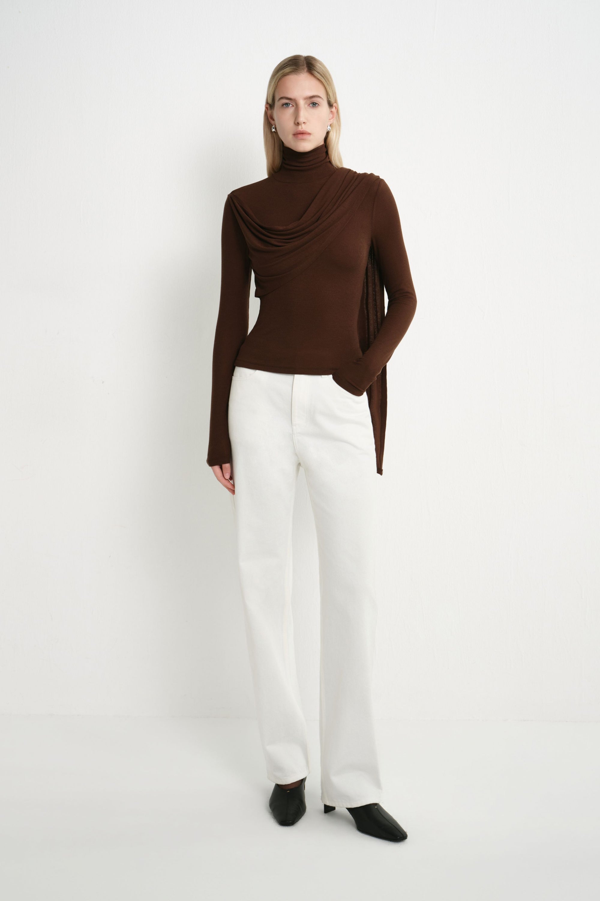 Front view of a model wearing a dark brown long-sleeve turtleneck top with asymmetric draping across the bodice, paired with white jeans and black shoes.