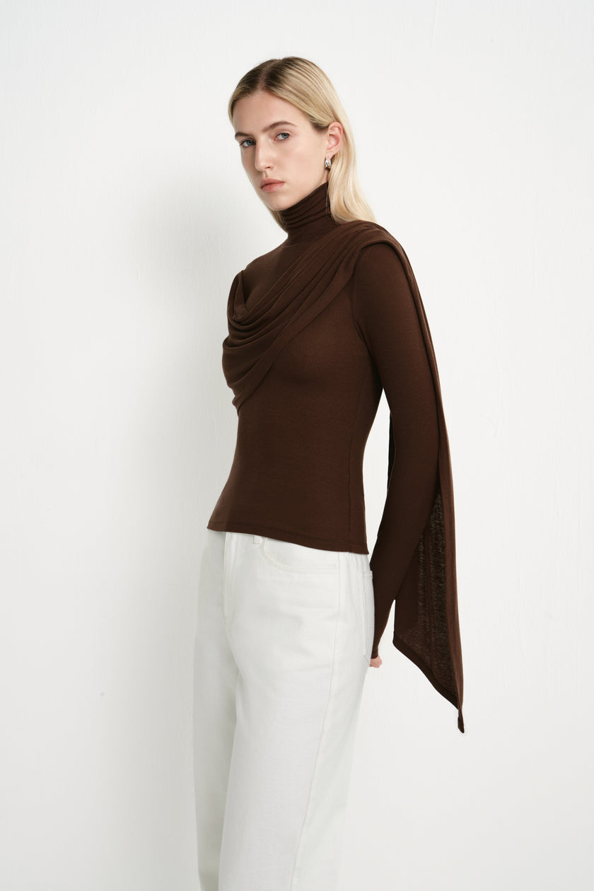 Three-quarter close-up of a dark brown turtleneck top featuring layered draping across the chest and an elongated asymmetric panel over one shoulder.