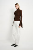 A model wearing a fitted dark brown turtleneck top with layered asymmetric draping and a long trailing side panel, styled with white straight-leg jeans.