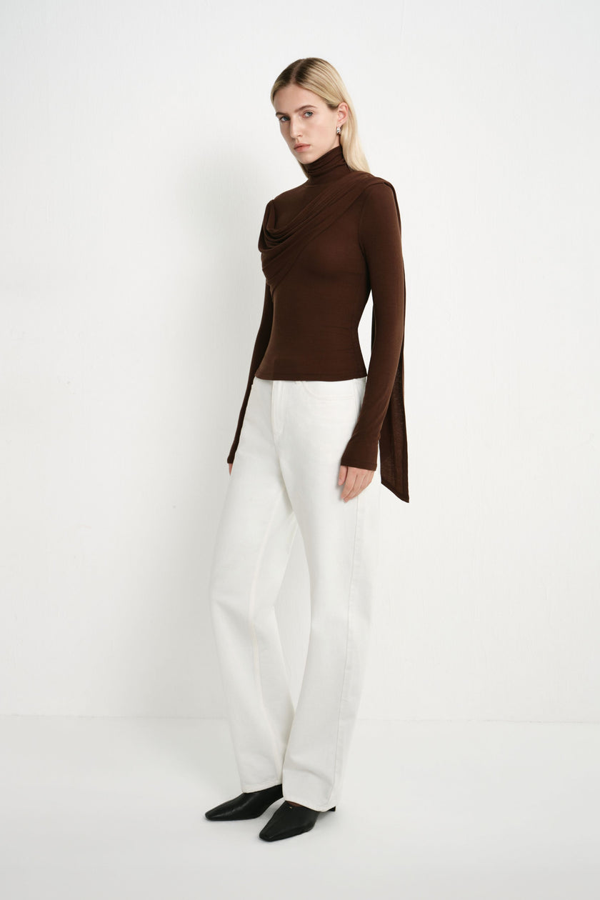 A model wearing a fitted dark brown turtleneck top with layered asymmetric draping and a long trailing side panel, styled with white straight-leg jeans.
