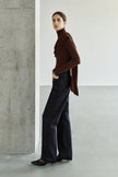 A model wearing a dark brown turtleneck top with asymmetric draping across the chest and a long scarf-like panel, styled with dark wide-leg jeans and black pointed shoes.
