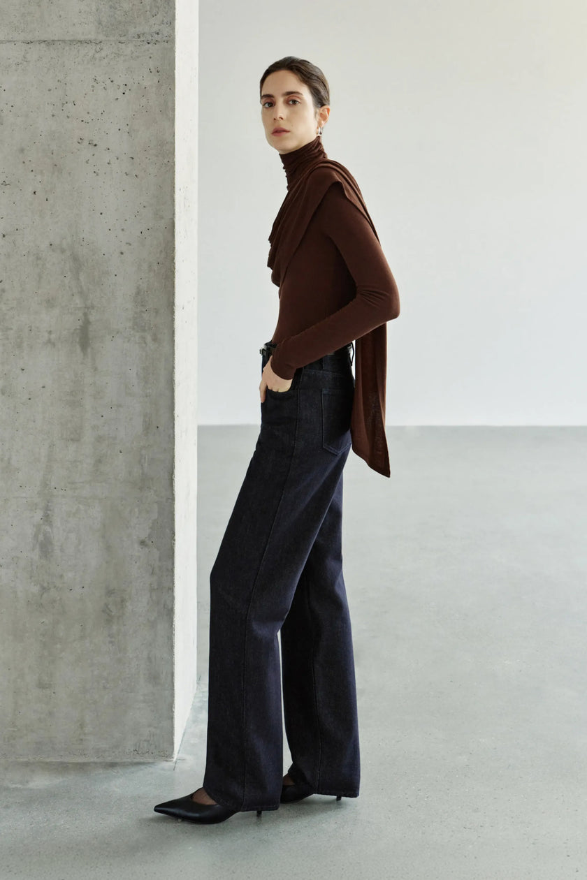 A model wearing a dark brown turtleneck top with asymmetric draping across the chest and a long scarf-like panel, styled with dark wide-leg jeans and black pointed shoes.