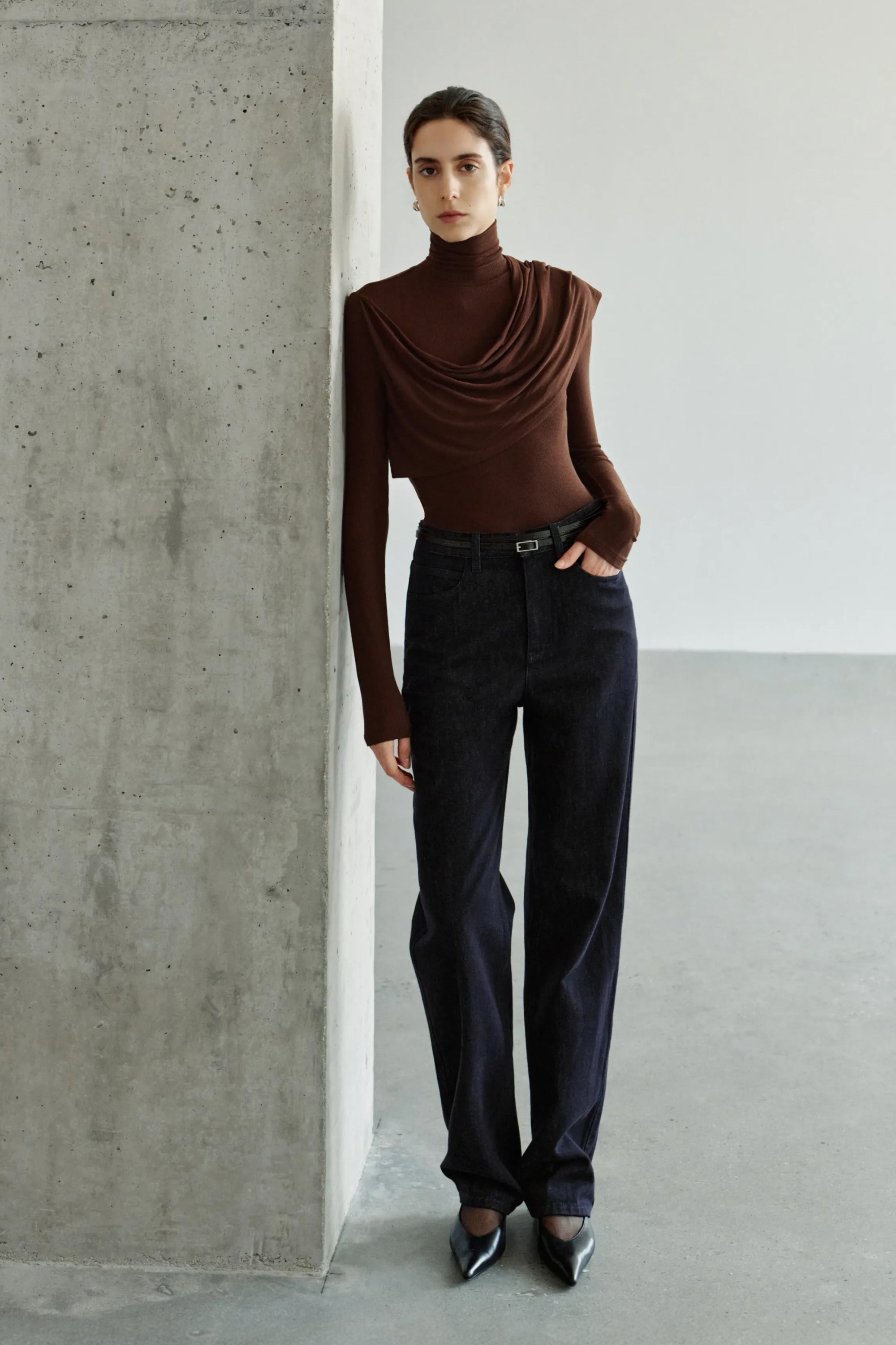 A model wearing a dark brown draped turtleneck top with an extended asymmetric shoulder panel, styled with a light beige A-line midi skirt and black pointed pumps.