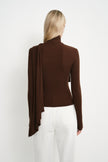 Back view of a dark brown fitted turtleneck top showing the long draped panel falling from one shoulder over the back, paired with white jeans.