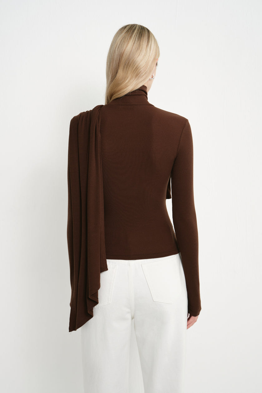 Back view of a dark brown fitted turtleneck top showing the long draped panel falling from one shoulder over the back, paired with white jeans.