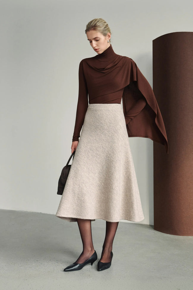 A model wearing a dark brown draped turtleneck top with an extended asymmetric shoulder panel, styled with a light beige A-line midi skirt and black pointed pumps.